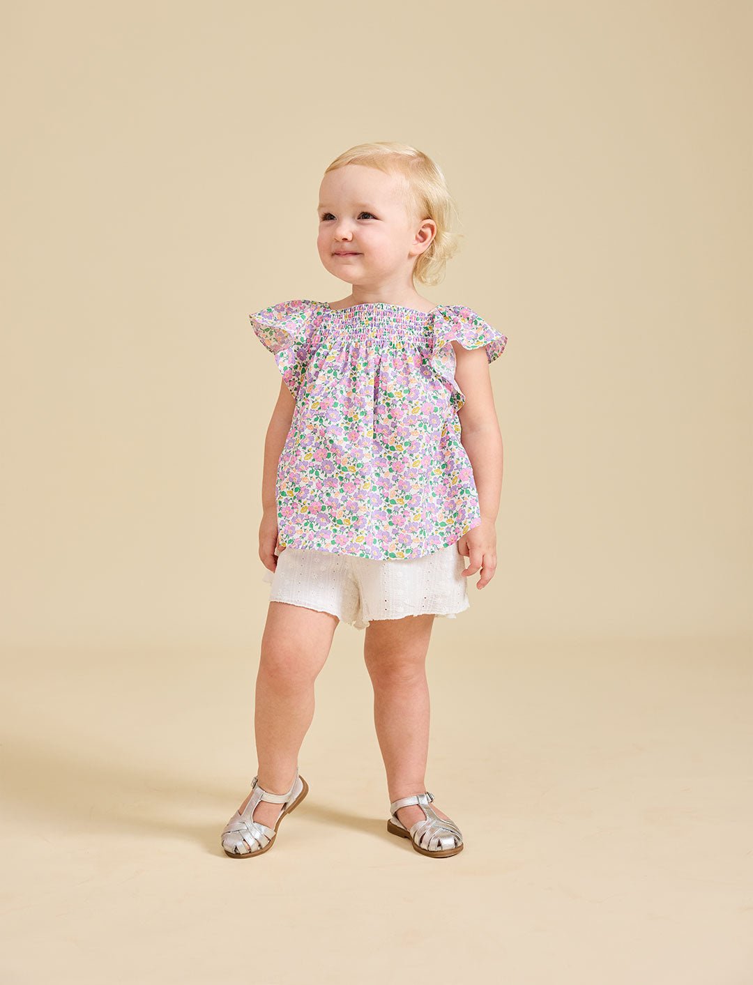 A toddler wearing a floral top and white shorts, standing against a beige background.