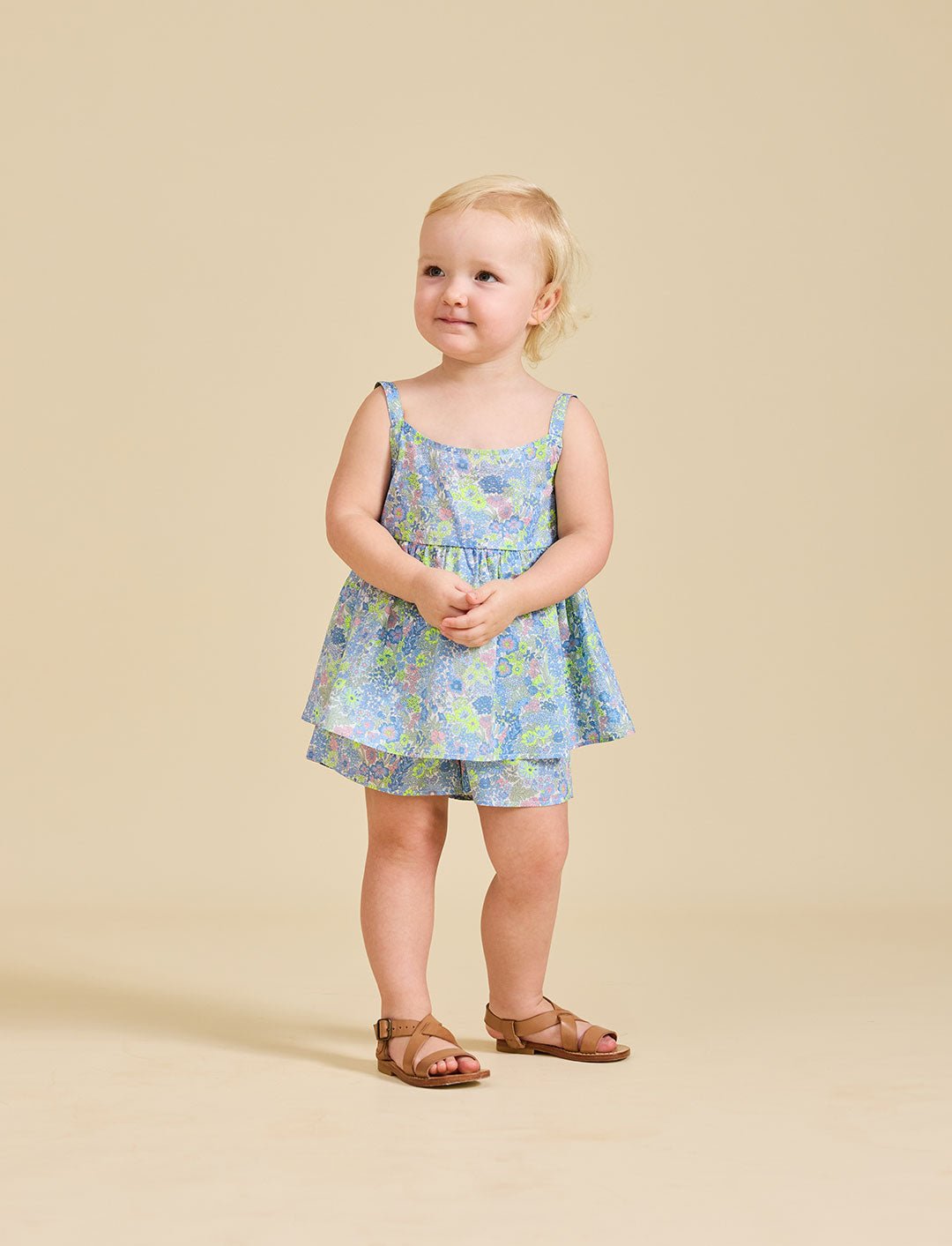 A young child standing against a beige background, wearing a floral dress with a sleeveless top and shorts, paired with sandals.