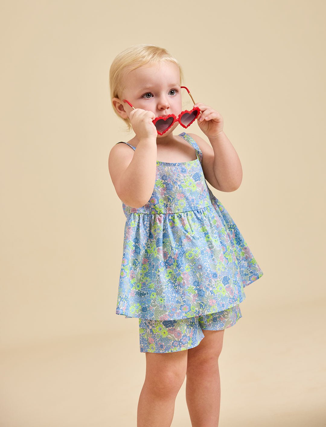 A child is standing, wearing a floral dress and shorts, holding sunglasses to their face.
