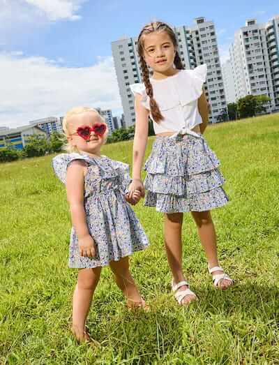 Two children in liberty print outfits playing in a grassy field with buildings in the background.
