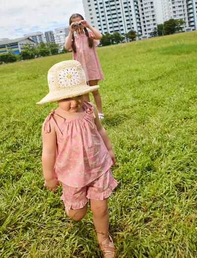 Two children in pink outfits playing in a grassy field with buildings in the background.