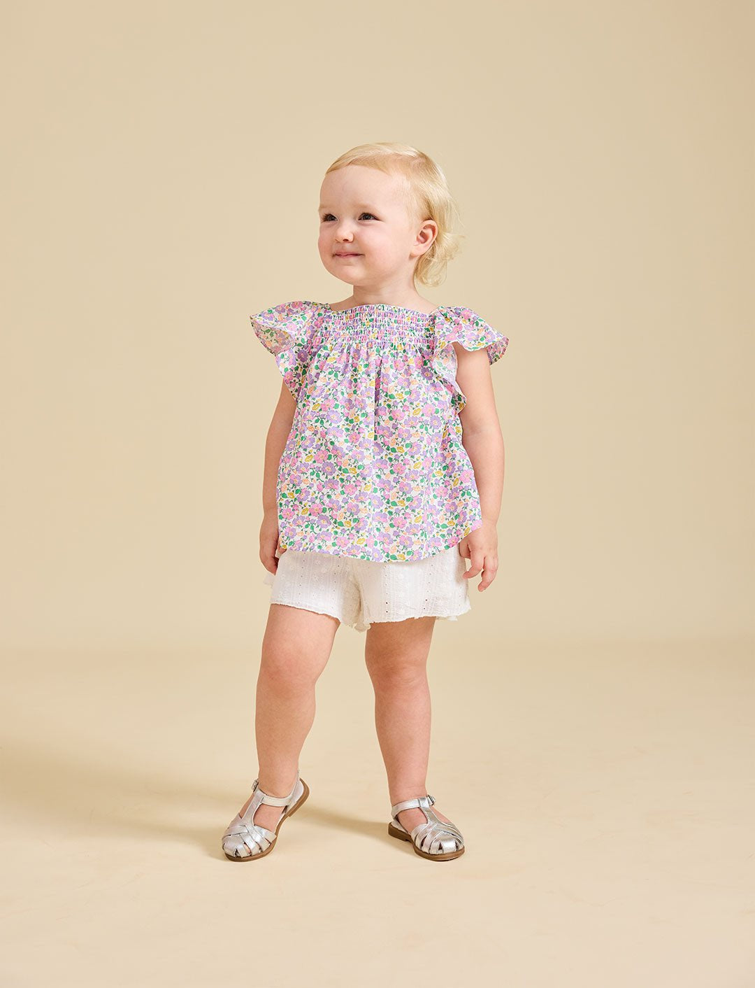 A toddler wearing a floral top and white shorts, standing against a beige background.