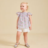A toddler wearing a floral top and white shorts, standing against a beige background.