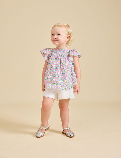 A toddler wearing a floral top and white shorts, standing against a beige background.