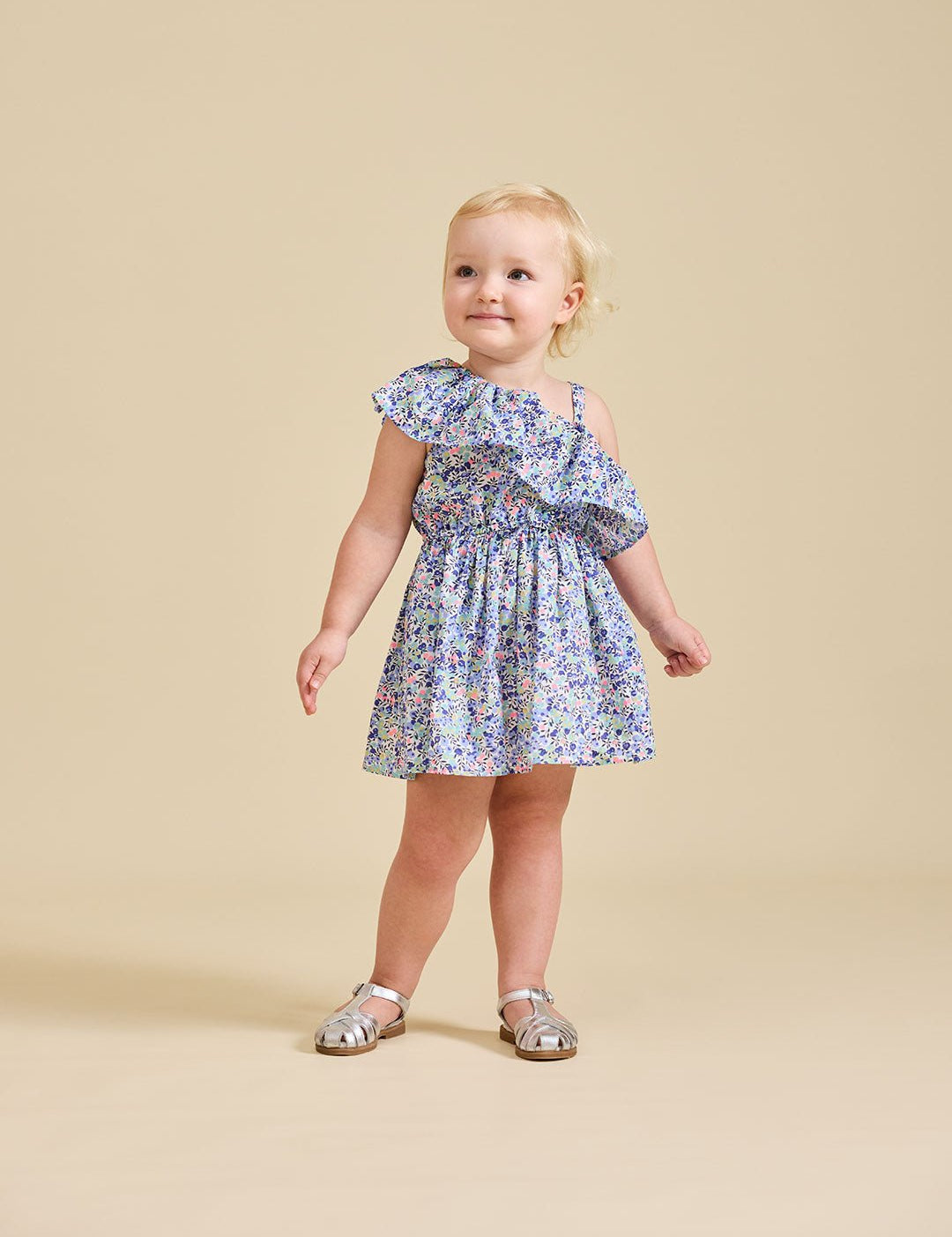 A young child standing against a beige background, wearing a floral dress with asymmetrical shoulder and frills, paired with white sandals.