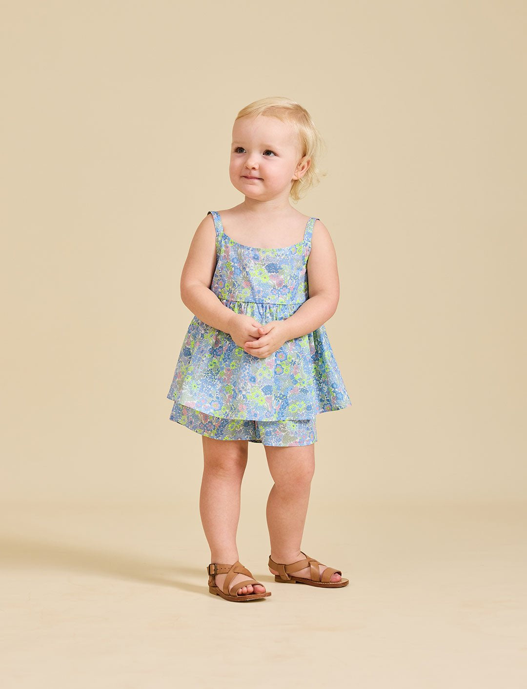 A young child standing against a beige background, wearing a floral dress with a sleeveless top and shorts, paired with sandals.