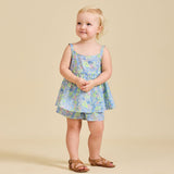A young child standing against a beige background, wearing a floral dress with a sleeveless top and shorts, paired with sandals.
