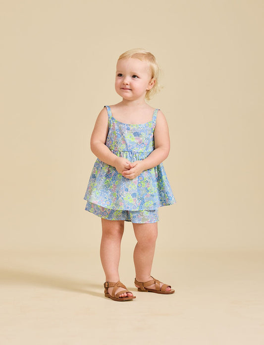 A young child standing against a beige background, wearing a floral dress with a sleeveless top and shorts, paired with sandals.
