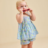 A child is standing, wearing a floral dress and shorts, holding sunglasses to their face.