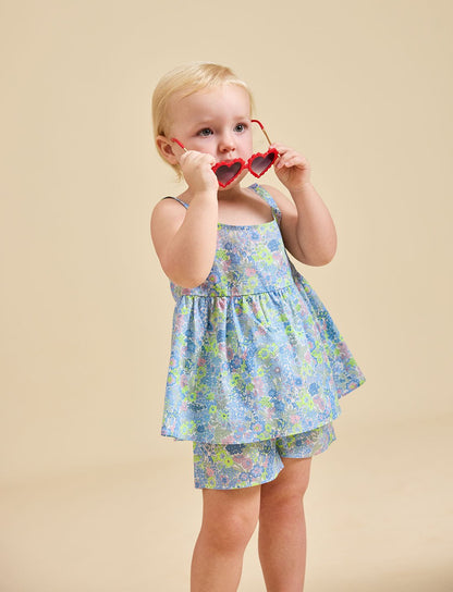 A child is standing, wearing a floral dress and shorts, holding sunglasses to their face.