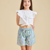 A girl wearing a white top with ruffled sleeves and blue floral shorts.