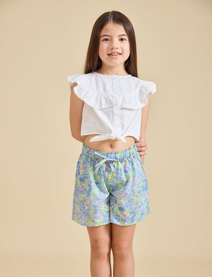 A girl wearing a white top with ruffled sleeves and blue floral shorts.