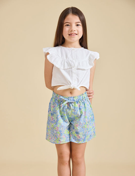 A girl wearing a white top with ruffled sleeves and blue floral shorts.