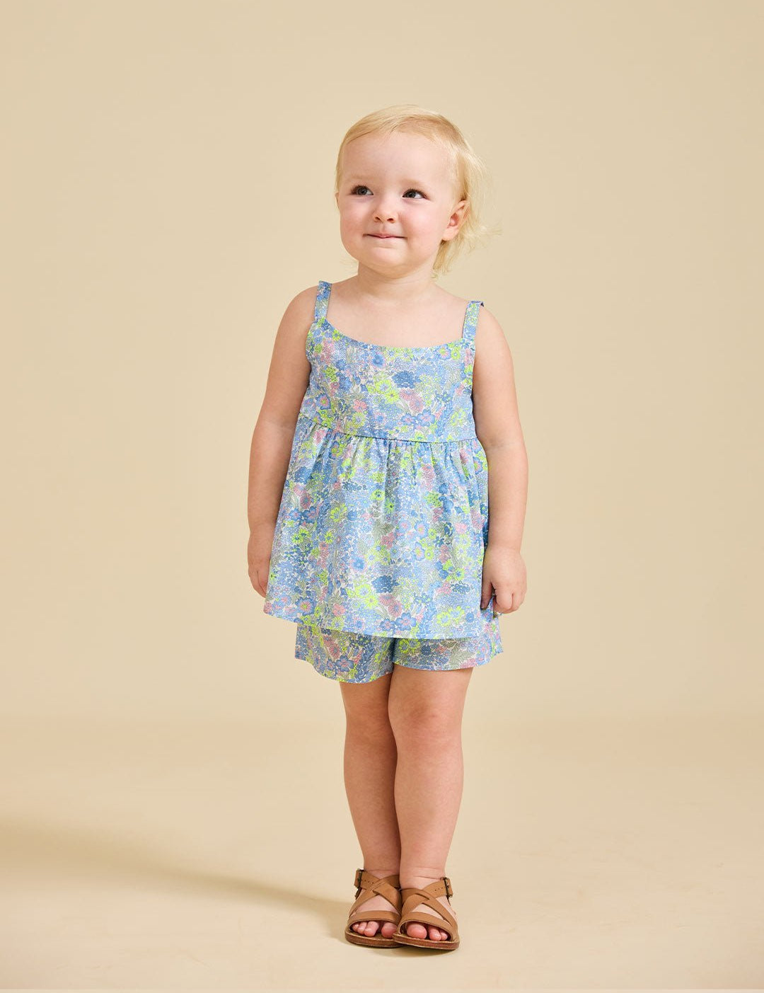A young child standing against a beige background, wearing a floral sleeveless top and shorts with a bow on the back.