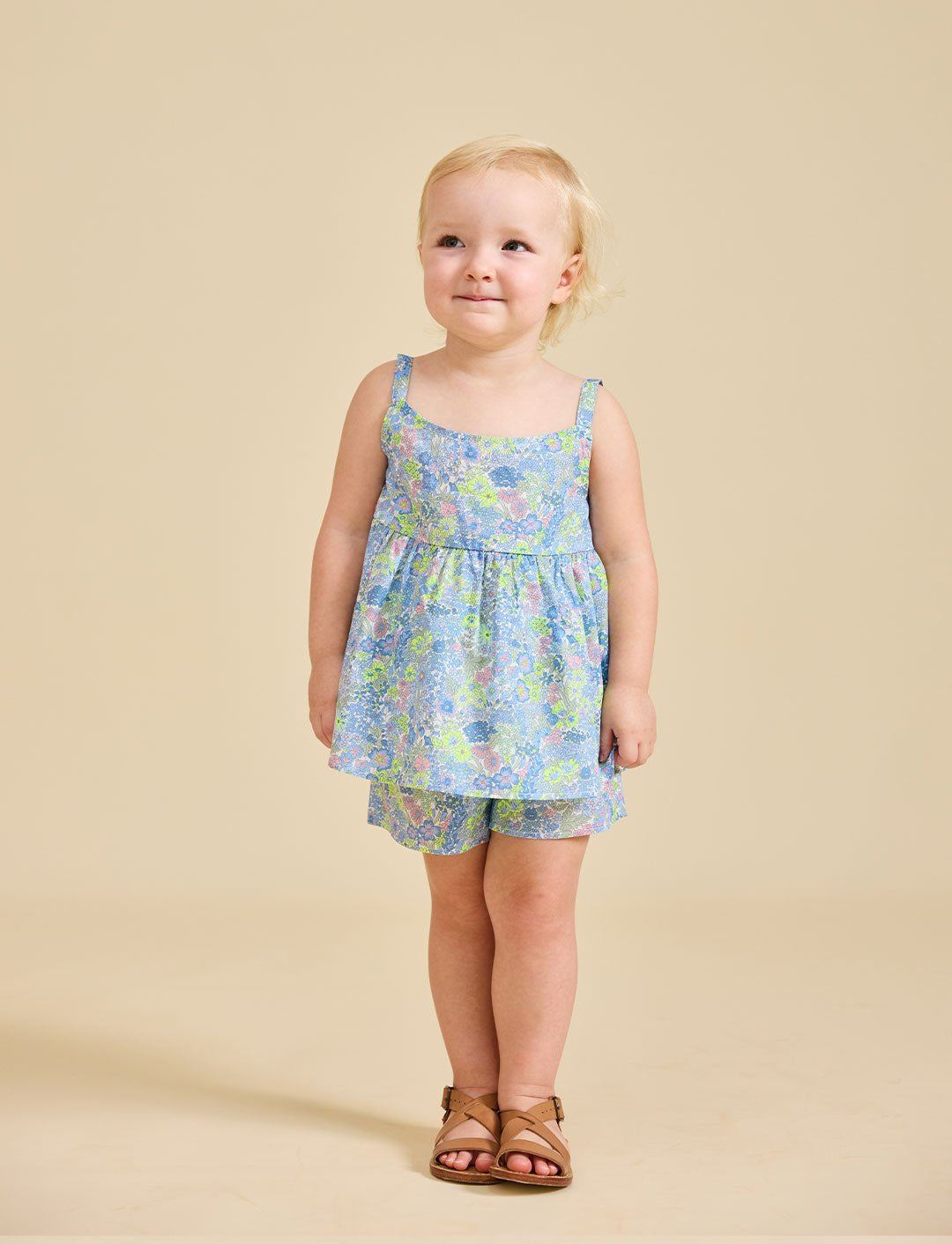 A young child standing against a beige background, wearing a floral sleeveless top and shorts with a bow on the back.