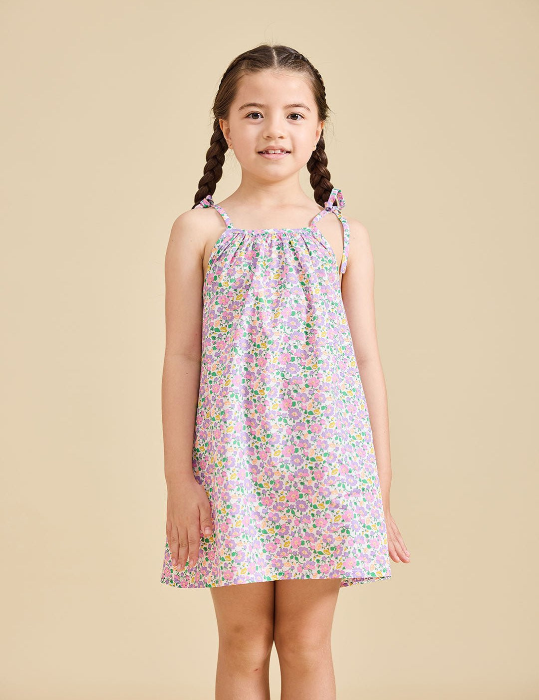 A young girl standing against a beige background, wearing the ce-jour la fleur sleeveless knee-length dress with adjustable shoulder straps.