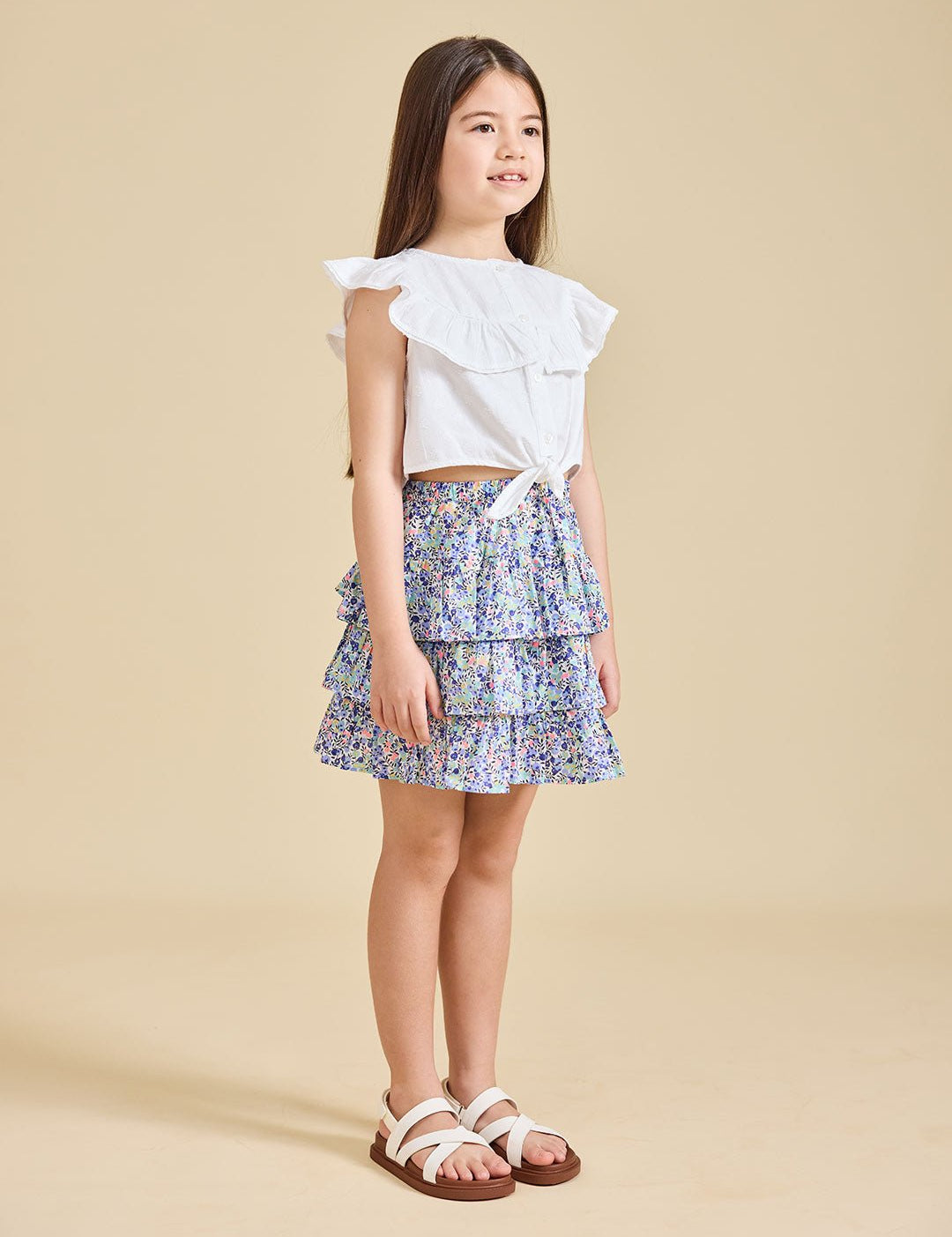 A young girl standing, wearing a white top with ruffled sleeves and a floral print Odette skirt with a pleated design. She is also wearing white sandals.