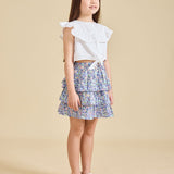 A young girl standing, wearing a white top with ruffled sleeves and a floral print Odette skirt with a pleated design. She is also wearing white sandals.