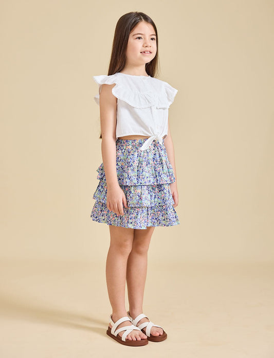 A young girl standing, wearing a white top with ruffled sleeves and a floral print Odette skirt with a pleated design. She is also wearing white sandals.