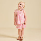 A young child standing against a beige background, wearing pink floral shorts and a matching sleeveless top with frill detailing.