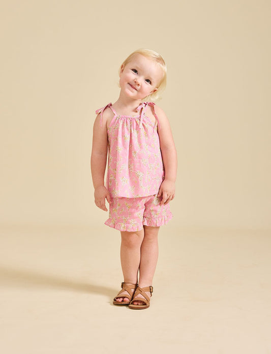 A young child standing against a beige background, wearing pink floral shorts and a matching sleeveless top with frill detailing.