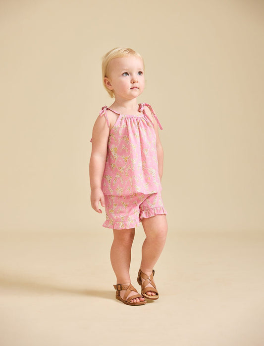 A child standing wearing a pink floral top and matching shorts with tan sandals.